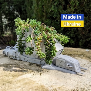 May include: A gray 3D printed planter shaped like a Star Wars AT-AT walker with green succulents growing out of the top. The planter has the text "Made in Ukraine" on a blue and yellow background.