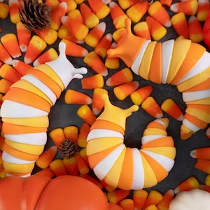 Puede incluir: Tres juguetes segmentados con forma de babosa en blanco, amarillo y naranja, rodeados de caramelos de maíz, calabazas y piñas. Los juguetes tienen un diseño lúdico, ideal para Halloween o la decoración de otoño.
