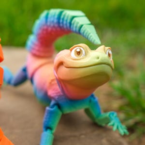 May include: A colorful, articulated lizard toy with a rainbow gradient of colors, including pink, blue, and green. The lizard has a friendly expression and is posed on a neutral surface. An orange, segmented object is in the foreground.
