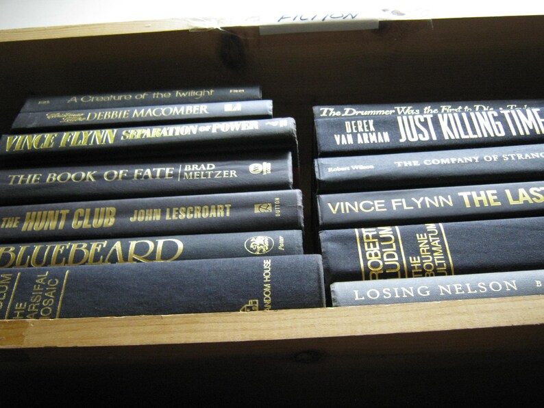 Puede incluir: Una estanter&iacute;a llena de libros de tapa dura negros con letras doradas. Los t&iacute;tulos incluyen "A Creature of the Twilight", "Separation of Power", "The Book of Fate", "The Hunt Club", "Bluebeard", "Just Killing Time", "The Company of Strange", "The Last", "The Bourne Ultimatum" y "Losing Nelson".