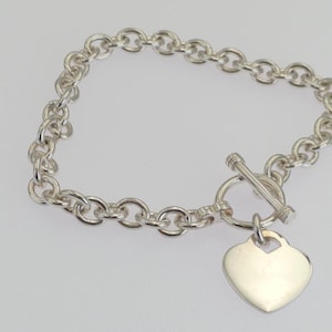 May include: A silver chain link bracelet featuring a toggle clasp closure and a smooth, heart-shaped pendant. The bracelet is constructed from polished silver-toned circular links, with the heart charm attached to the circular toggle ring.