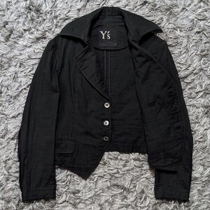 May include: A black blazer featuring a wide collar, long sleeves, and a three-button front closure. The garment includes a small flap pocket on the lower front and a label at the inner neckline with the text Y's printed in white lettering.