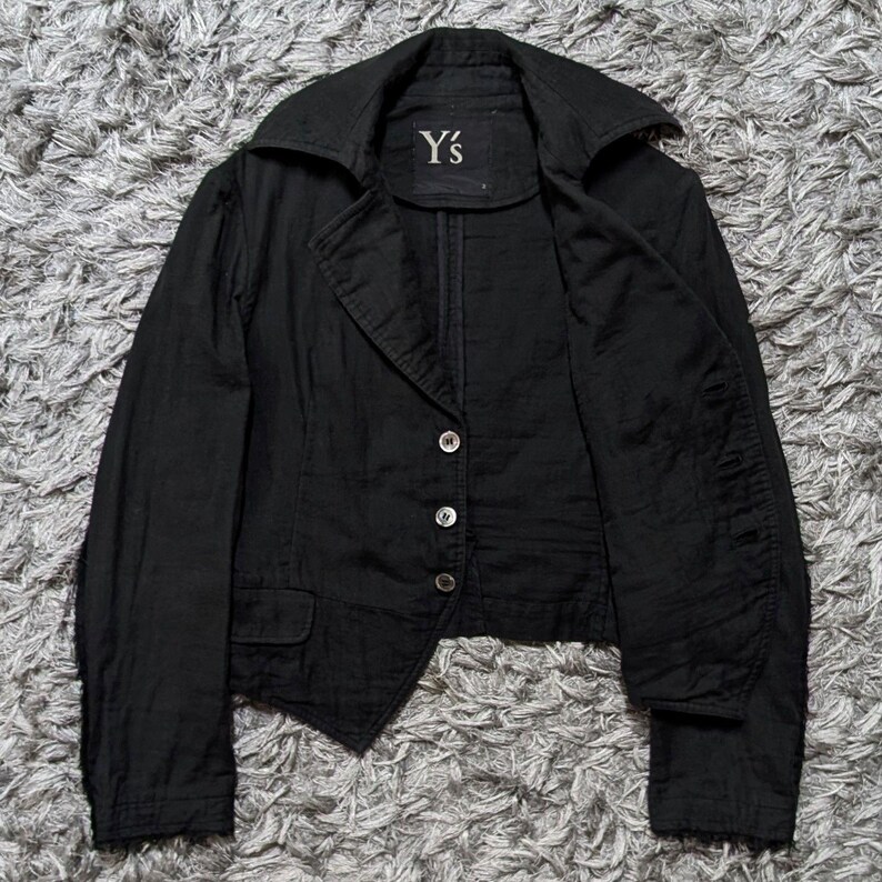 May include: A black blazer featuring a wide collar, long sleeves, and a three-button front closure. The garment includes a small flap pocket on the lower front and a label at the inner neckline with the text Y's printed in white lettering.