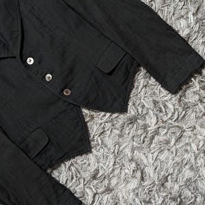 May include: A black blazer featuring a notched lapel, long sleeves, and a front closure with three silver-toned buttons. The garment includes two front flap pockets and a pointed hemline, constructed with visible stitching along the seams.