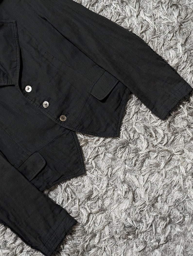 May include: A black blazer featuring a notched lapel, long sleeves, and a front closure with three silver-toned buttons. The garment includes two front flap pockets and a pointed hemline, constructed with visible stitching along the seams.