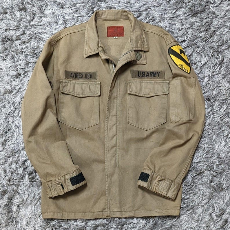 May include: A tan long-sleeve jacket featuring a fold-down collar, a front zip fastening, and two chest pockets. The jacket displays embroidered patches reading AVIREX USA and U.S. ARMY, along with a yellow and black shoulder patch emblem.