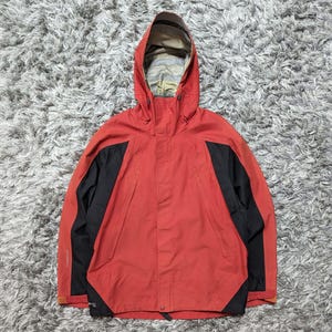May include: A red hooded jacket with black side panels. The jacket features a full zip and a hood with a patterned lining. The jacket is made from a durable, weather-resistant material, suitable for outdoor activities.