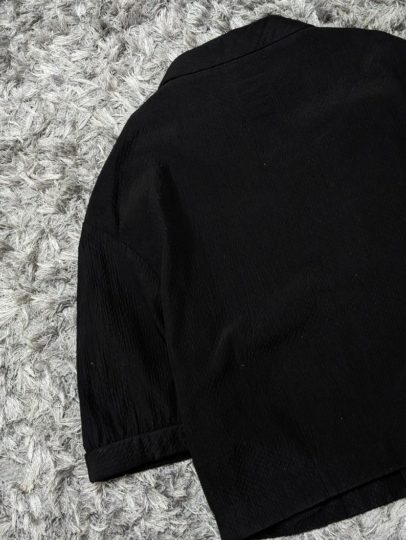 May include: A solid black blazer featuring a textured fabric with vertical ribbed detailing, a notched lapel collar, and three-quarter length sleeves with cuffed hems. The garment is constructed with a structured shoulder design and a clean finish.
