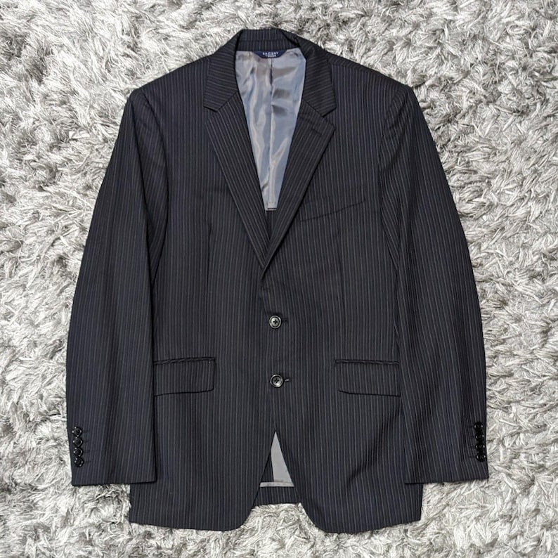 May include: A dark gray pinstripe blazer with a two-button closure. The blazer has a notched lapel, a chest pocket, and two front pockets. The sleeves have four buttons. The interior lining is a light gray color.