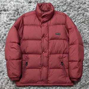 May include: A brick red puffer jacket featuring a high collar, a front button placket, and two zippered side pockets. The jacket has a horizontal quilted pattern and a small rectangular black patch on the chest with the white text Converse.