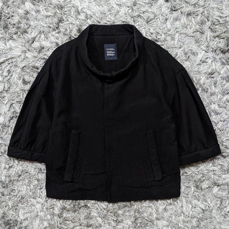May include: A black jacket with a high collar, three-quarter length sleeves, and two vertical front pockets. The garment features a textured fabric surface and a dark interior label with the white text LIMI feu visible at the neckline.