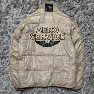 Peut inclure: Veste bouffante beige avec le texte "AERO SERVICE" et un embl&egrave;me ail&eacute; noir et blanc au dos. La veste a un col montant et un design matelass&eacute;. La veste est faite d'un mat&eacute;riau brillant.