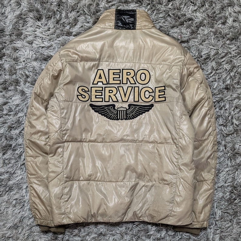 Peut inclure: Veste bouffante beige avec le texte "AERO SERVICE" et un embl&egrave;me ail&eacute; noir et blanc au dos. La veste a un col montant et un design matelass&eacute;. La veste est faite d'un mat&eacute;riau brillant.