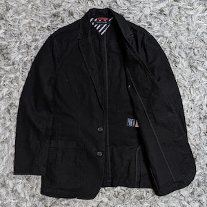 May include: A black blazer featuring a two-button front closure, two lower patch pockets, and a notched lapel. The interior lining displays a white and navy blue striped pattern with a red accent at the collar, and a Tommy Hilfiger brand label.