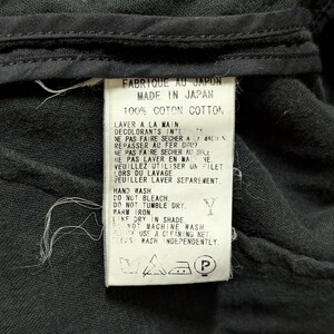 May include: A white rectangular garment care label sewn onto black fabric. The label features black text in French and English, stating Made in Japan, 100% Cotton, and provides washing instructions including hand wash, do not bleach, and line dry.