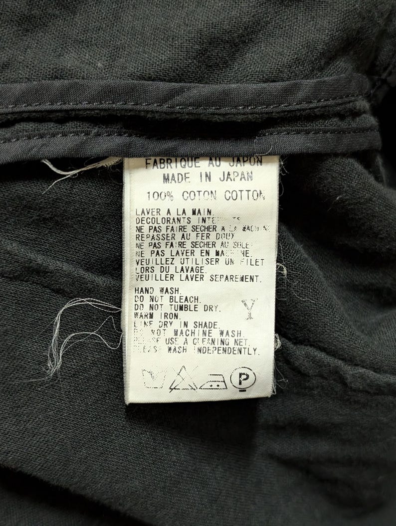 May include: A white rectangular garment care label sewn onto black fabric. The label features black text in French and English, stating Made in Japan, 100% Cotton, and provides washing instructions including hand wash, do not bleach, and line dry.