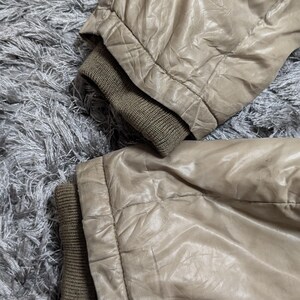 Peut inclure: Gros plan sur deux manches de veste beiges avec des poignets c&ocirc;tel&eacute;s vert olive. Le tissu de la veste a un aspect brillant et l&eacute;g&egrave;rement froiss&eacute;. Les manches reposent sur une surface grise textur&eacute;e.