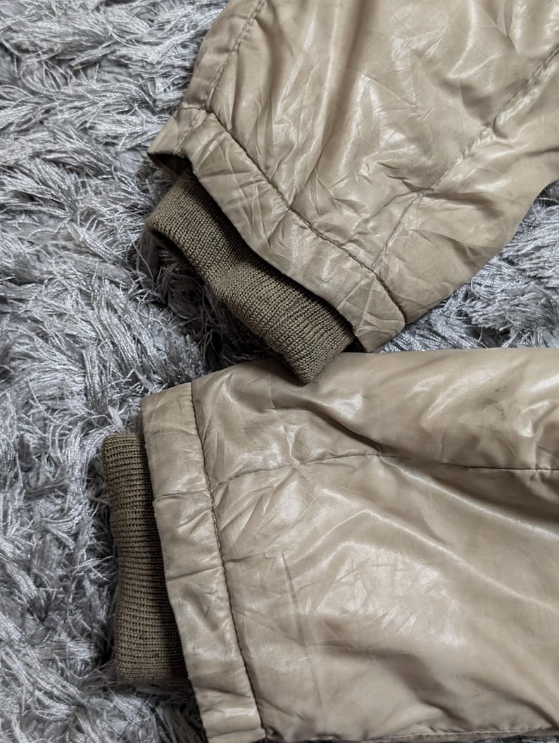 Peut inclure: Gros plan sur deux manches de veste beiges avec des poignets c&ocirc;tel&eacute;s vert olive. Le tissu de la veste a un aspect brillant et l&eacute;g&egrave;rement froiss&eacute;. Les manches reposent sur une surface grise textur&eacute;e.