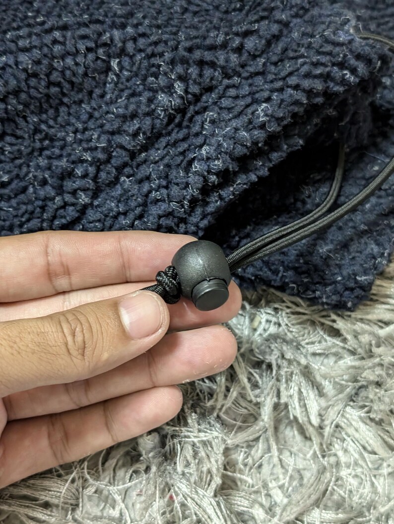 May include: Close-up of a navy blue fleece fabric with a drawstring and a black cord lock. The fabric has a textured, fuzzy appearance. The cord lock is round and made of black plastic.