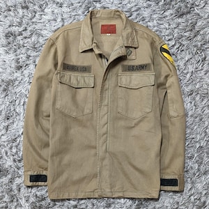 May include: A tan long-sleeve jacket with a collar, a front zip, and two chest pockets. The jacket features a red label on the inner collar, a black patch on the left sleeve, and two black embroidered name tapes reading AVIREX USA and U.S. ARMY.