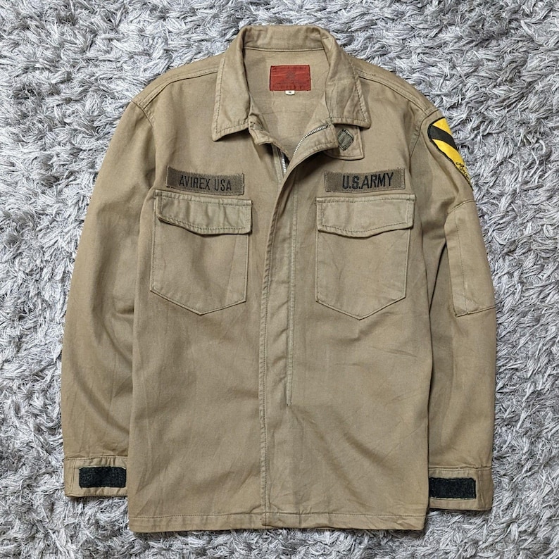May include: A tan long-sleeve jacket with a collar, a front zip, and two chest pockets. The jacket features a red label on the inner collar, a black patch on the left sleeve, and two black embroidered name tapes reading AVIREX USA and U.S. ARMY.