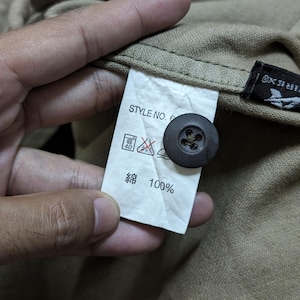 May include: A tan-coloured garment featuring a white interior care label with the text STYLE NO. 6 and 100% cotton in Japanese characters. A dark brown, four-hole button is attached to the fabric near the label, which also displays laundry symbols.