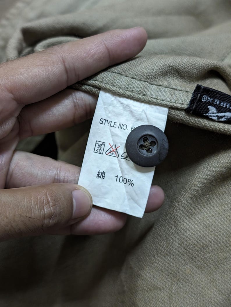 May include: A tan-coloured garment featuring a white interior care label with the text STYLE NO. 6 and 100% cotton in Japanese characters. A dark brown, four-hole button is attached to the fabric near the label, which also displays laundry symbols.
