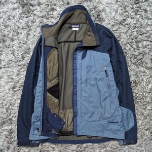 Könnte beinhalten: Eine marineblaue und hellblaue Patagonia Jacke mit durchgehendem Reißverschluss und mehreren Taschen. Die Jacke hat einen hohen Kragen und ein Mesh-Futter. Das Patagonia-Logo ist auf der Brust sichtbar. Die Jacke ist offen und zeigt das Innere.