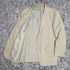 May include: A beige blazer featuring a notched lapel, a three-button front closure, and two lower patch pockets with flaps. The interior is partially lined in a matching beige fabric with a red trim detail and a white rectangular brand label.