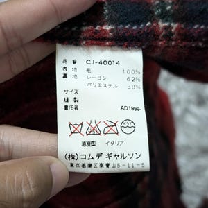 May include: Close-up of a tag with Japanese text and care symbols. The tag is attached to a red, black, and green plaid fabric. The tag includes the text "CJ-40014" and "AD1999-".