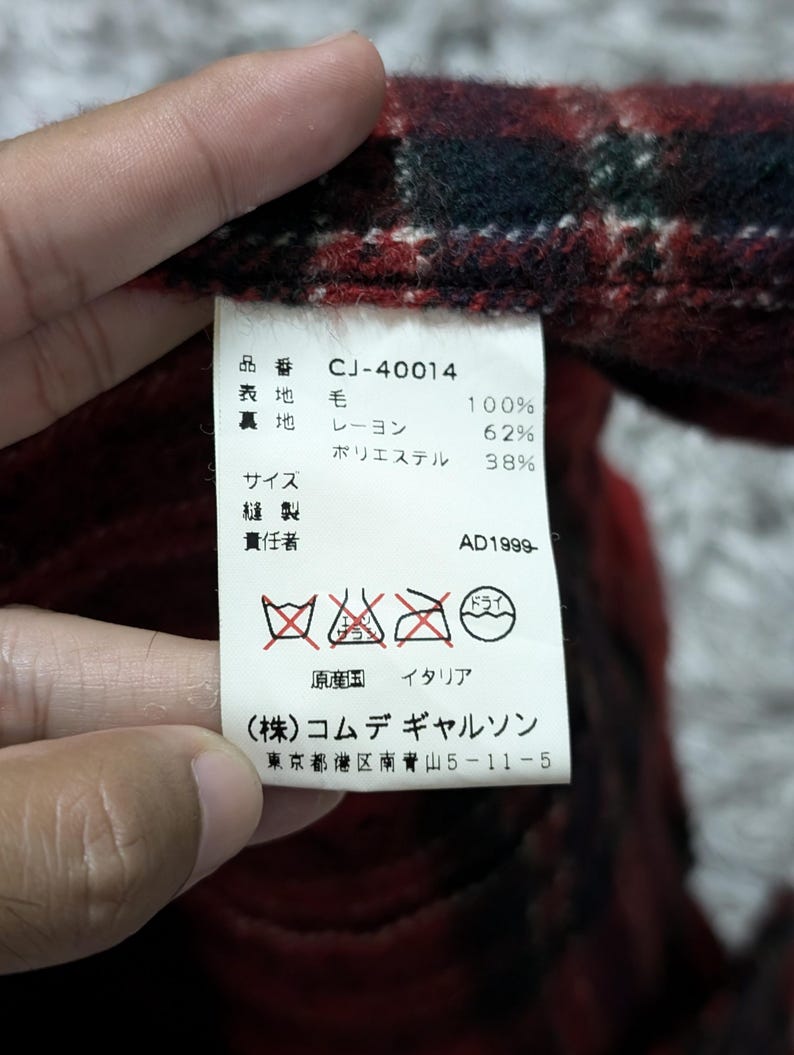 May include: Close-up of a tag with Japanese text and care symbols. The tag is attached to a red, black, and green plaid fabric. The tag includes the text "CJ-40014" and "AD1999-".