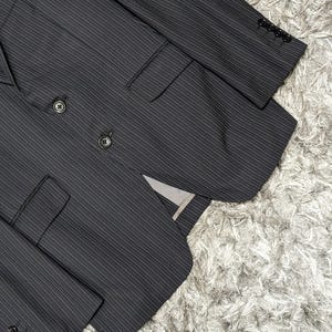 May include: A dark gray pinstripe suit jacket with a double-breasted design. The jacket features two rows of buttons, a notched lapel, and multiple pockets. The sleeves have buttons at the cuffs. The suit is made of a textured fabric.