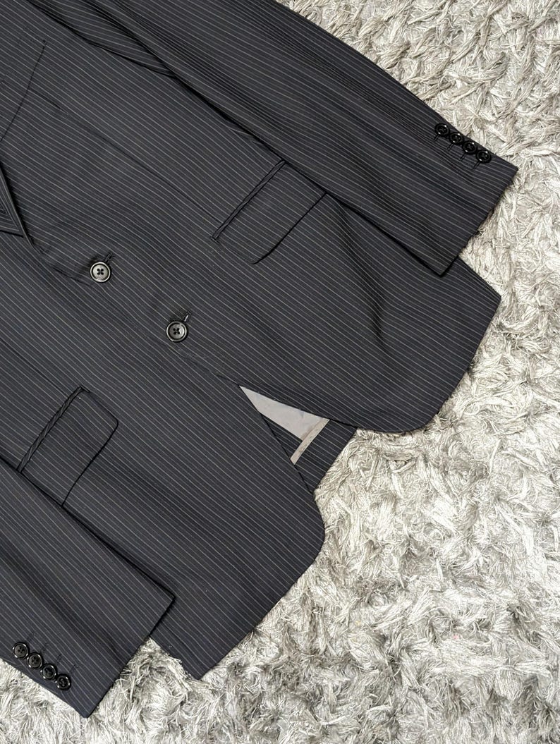 May include: A dark gray pinstripe suit jacket with a double-breasted design. The jacket features two rows of buttons, a notched lapel, and multiple pockets. The sleeves have buttons at the cuffs. The suit is made of a textured fabric.
