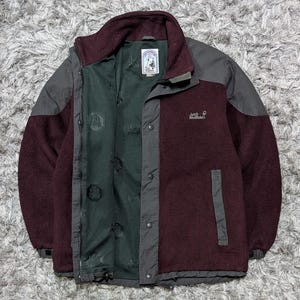 May include: A burgundy and gray fleece jacket with a high collar and a full zipper. The jacket features a dark green interior lining and the brand name "Jack Wolfskin" embroidered on the chest. The jacket has two side pockets and a small logo patch.