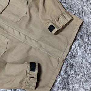 May include: A tan utility jacket featuring a structured collar, two chest pockets with flaps, and long sleeves with adjustable cuffs secured by black hook-and-loop fasteners. The garment is constructed from a durable, solid-coloured fabric.