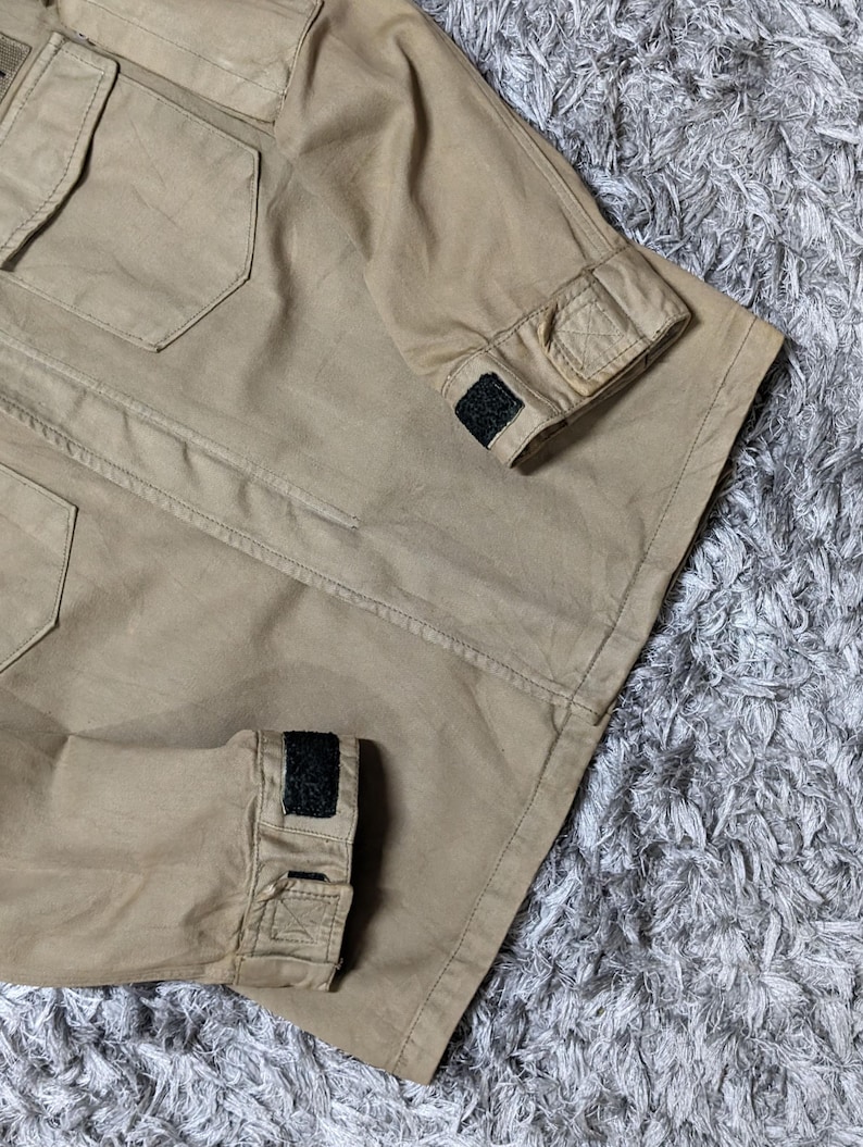 May include: A tan utility jacket featuring a structured collar, two chest pockets with flaps, and long sleeves with adjustable cuffs secured by black hook-and-loop fasteners. The garment is constructed from a durable, solid-coloured fabric.