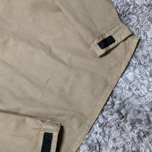 May include: A tan long-sleeved jacket featuring a straight hemline and cuffs that are adjustable with black hook-and-loop fasteners. The garment is constructed from a solid-coloured fabric with visible stitching along the seams and sleeve edges.