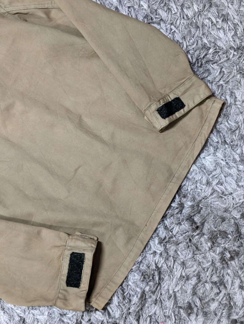 May include: A tan long-sleeved jacket featuring a straight hemline and cuffs that are adjustable with black hook-and-loop fasteners. The garment is constructed from a solid-coloured fabric with visible stitching along the seams and sleeve edges.