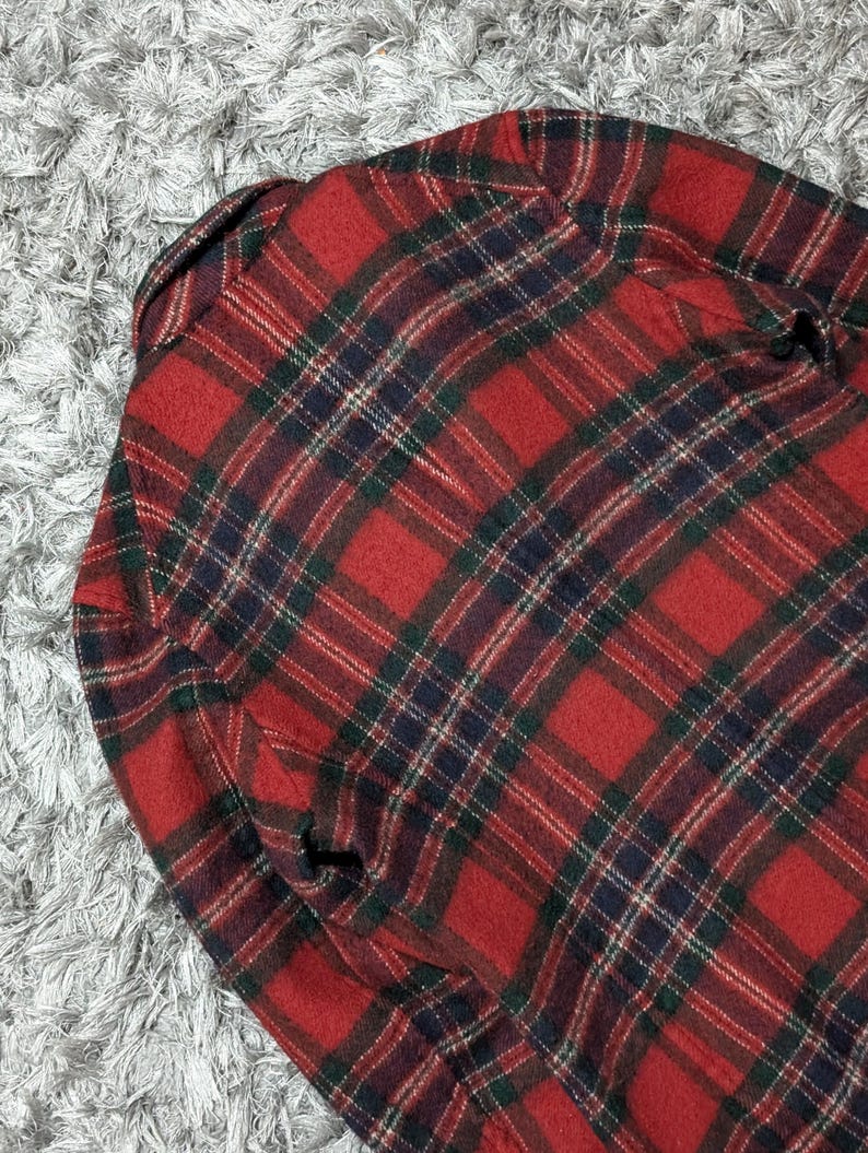 May include: A red and black plaid flannel shirt. The shirt features a classic plaid pattern with green and navy blue accents. The fabric appears to be a soft, woven material, and the shirt is folded.