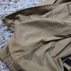 May include: A tan-coloured long-sleeved shirt made of a durable, woven fabric. The garment features a structured collar, a visible chest pocket with a flap, and a strip of black hook-and-loop fastener tape on the sleeve cuff area.