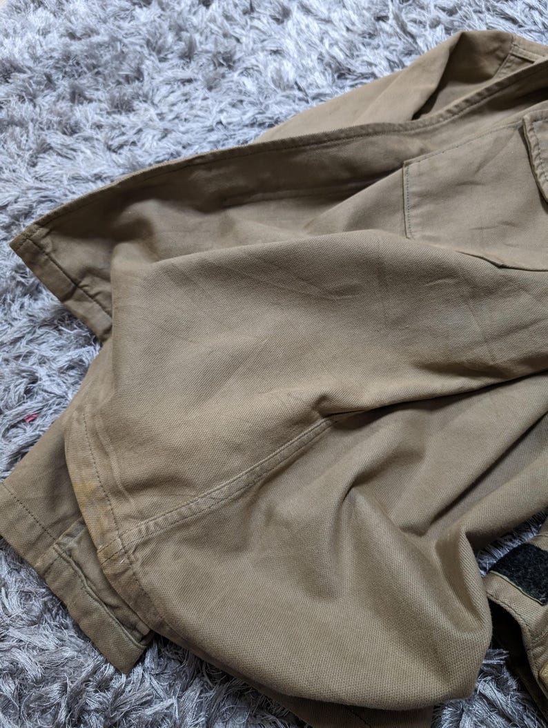 May include: A tan-coloured long-sleeved shirt made of a durable, woven fabric. The garment features a structured collar, a visible chest pocket with a flap, and a strip of black hook-and-loop fastener tape on the sleeve cuff area.