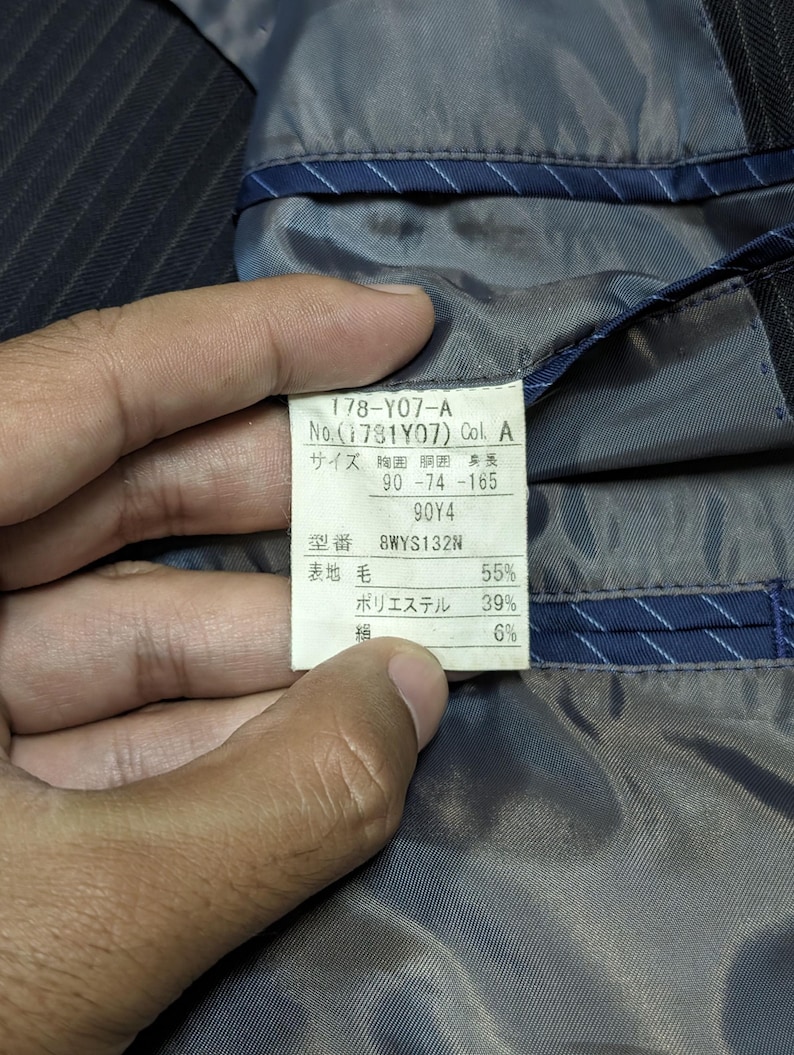 May include: Close-up of a label inside a dark pinstripe suit jacket. The label displays text in Japanese, including size and material composition percentages: 55% wool, 39% polyester, and 6% other.
