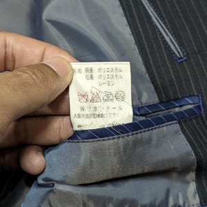 May include: Close-up of a dark blue pinstripe suit jacket interior. A tag with Japanese text is visible, along with care symbols. The lining is a light blue-gray color, with blue trim and stitching. The jacket appears to be well-tailored.