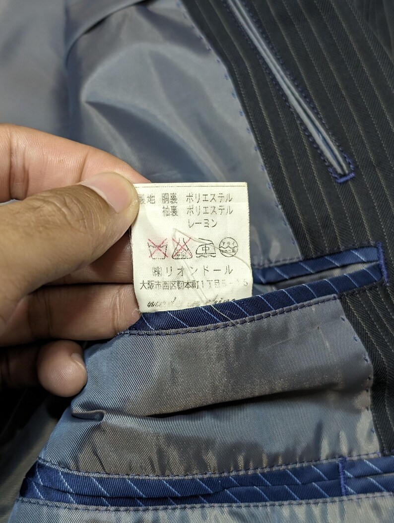 May include: Close-up of a dark blue pinstripe suit jacket interior. A tag with Japanese text is visible, along with care symbols. The lining is a light blue-gray color, with blue trim and stitching. The jacket appears to be well-tailored.