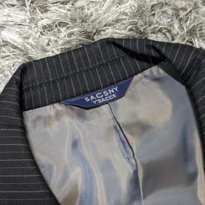 May include: Close-up of a black pinstripe blazer with a gray lining. The blazer's label reads "SACSNY Y'SACCS" in white text on a navy blue background. The fabric has a subtle sheen.