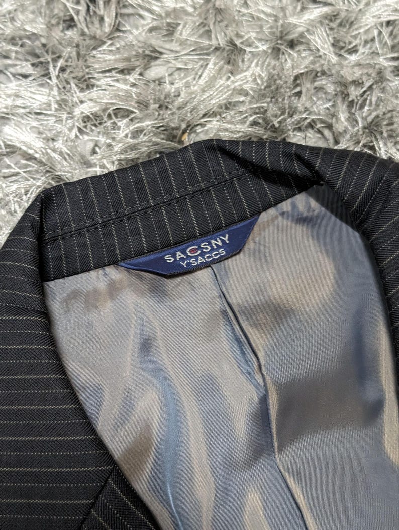 May include: Close-up of a black pinstripe blazer with a gray lining. The blazer's label reads "SACSNY Y'SACCS" in white text on a navy blue background. The fabric has a subtle sheen.