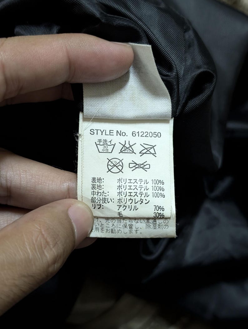 Peut inclure: Gros plan sur une &eacute;tiquette de v&ecirc;tement avec le texte "STYLE No. 6122050". L'&eacute;tiquette indique la composition du tissu : 100% polyester pour les couches ext&eacute;rieure et int&eacute;rieure, 70% acrylique et 30% laine pour les bord-c&ocirc;tes. Instructions de lavage incluses.