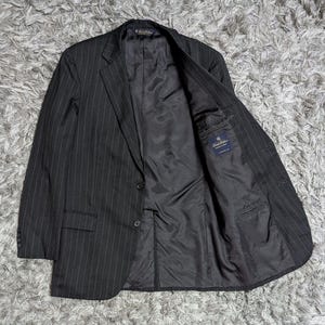 Peut inclure: Un blazer gris anthracite à fines rayures avec un revers cranté, deux boutons sur le devant et quatre boutons sur chaque poignet. L'intérieur est entièrement doublé de tissu noir et comporte une étiquette Brooks Brothers dans la poche.