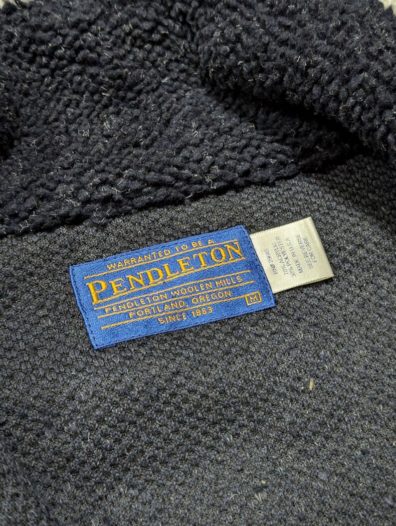 May include: Close-up of a dark navy blue Pendleton jacket. The blue label reads "PENDLETON WOOLEN MILLS PORTLAND, OREGON SINCE 1863". The jacket has a textured, fuzzy interior and a woven exterior. A white tag with care instructions is also visible.