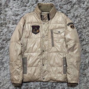 Peut inclure: Veste matelass&eacute;e beige avec un col montant, fermeture &eacute;clair et boutons. La veste comporte plusieurs poches et &eacute;cussons avec du texte et des embl&egrave;mes. La veste est de couleur neutre et semble &ecirc;tre faite d'un mat&eacute;riau brillant.